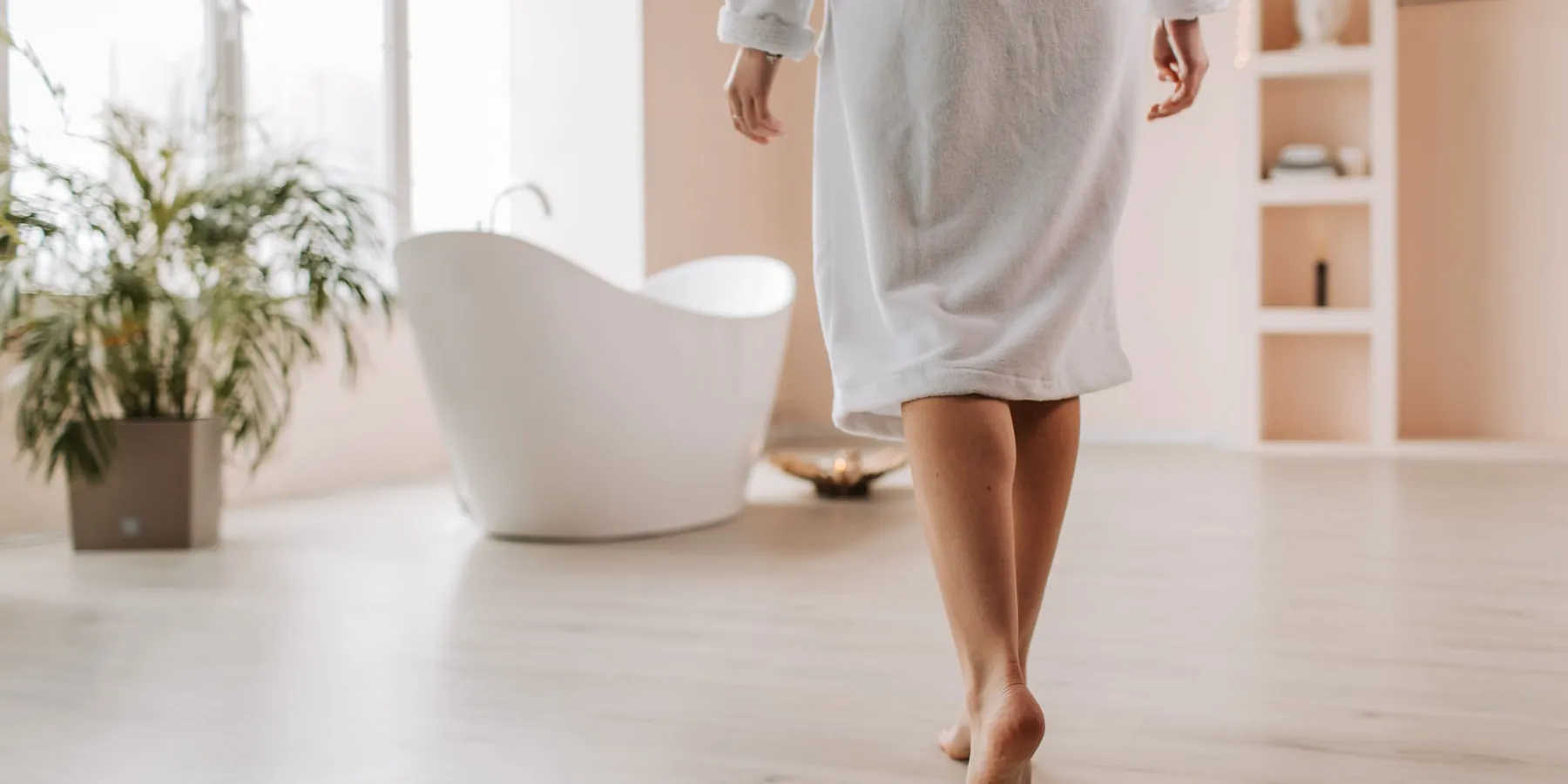 Une femme en peignoir blanc marche vers une baignoire, lumière du jour